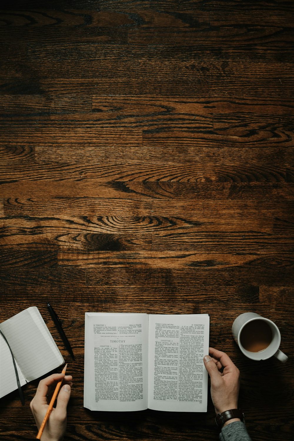 Hands resting on an open Bible beside a notebook and a cup of coffee on a wooden table